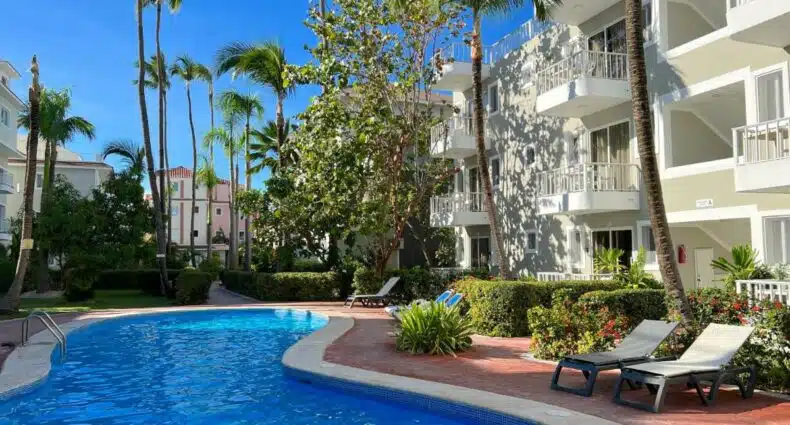 Área da piscina das Sol Caribe Suites, um dos hotéis em Punta Cana. A piscina em zigue-zague é rodeada por espreguiçadeiras no lado direito. Há várias árvores altas ao fundo, e o hotel com fachada branca fica atrás das espreguiçadeiras. O céu azul está límpido acima.