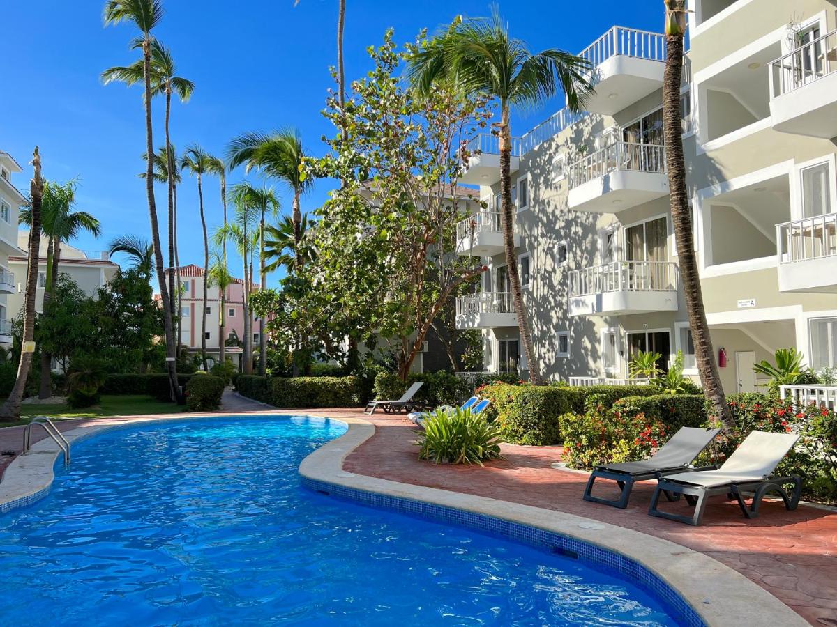 Área da piscina das Sol Caribe Suites, um dos hotéis em Punta Cana. A piscina em zigue-zague é rodeada por espreguiçadeiras no lado direito. Há várias árvores altas ao fundo, e o hotel com fachada branca fica atrás das espreguiçadeiras. O céu azul está límpido acima.