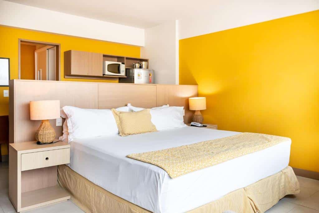 Quarto no Sotero Hotel, ilustrando o post de hotéis para o carnaval de Salvador. A cama está no centro da foto, e ela é box de casal. Em ambos seus lados, há uma mesa de cabeceira com abajur e gaveta. Atrás da cabeceira da cama, podemos ver de relance a cozinha da suíte.