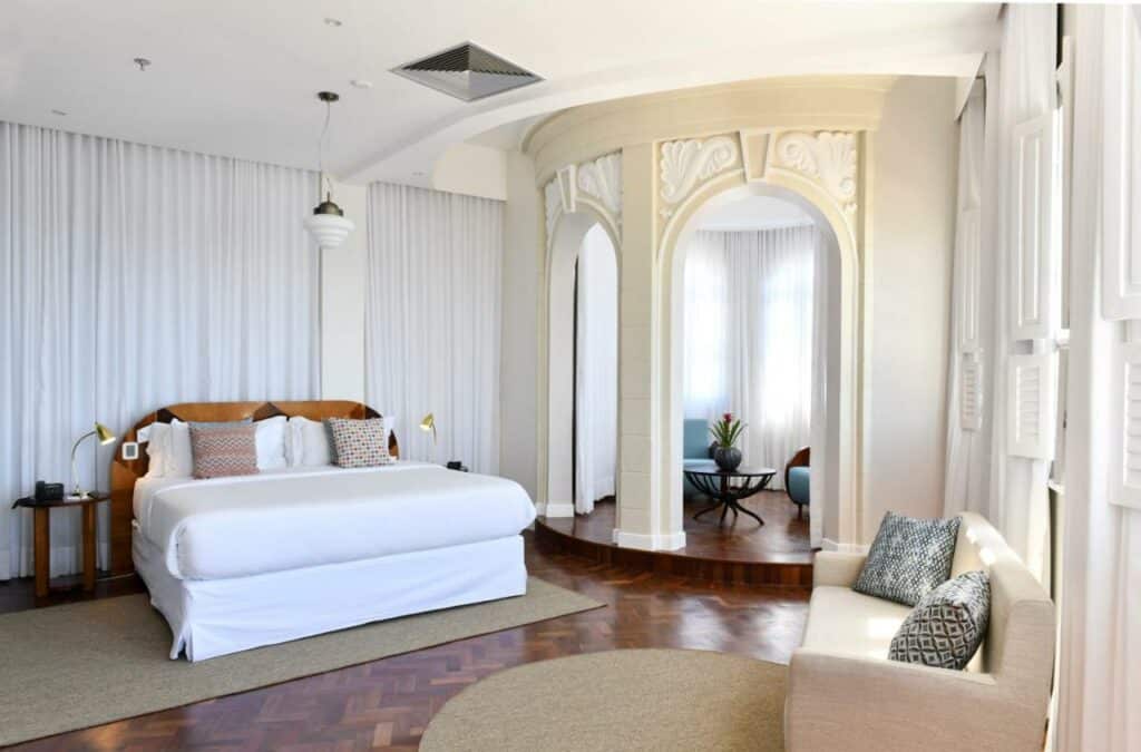 Uma suíte no Fera Palace Hotel, ilustrando o post de hotéis para o carnaval de Salvador. O ambiente é extremamente elegante, com decorações clássicas e o branco predomina. A cama é king-sized, e em ambos os lados há uma mesa de cabeceira com um abajur. Já no canto direito da foto, há um sofá rose com almofadas, perto das janelas. Mais ao fundo do quarto, do lado direito da cama, há uma plataforma com um arco clássico, e nele há poltronas e uma mesa de centro. Todo o quarto é rodeado por cortinas brancas