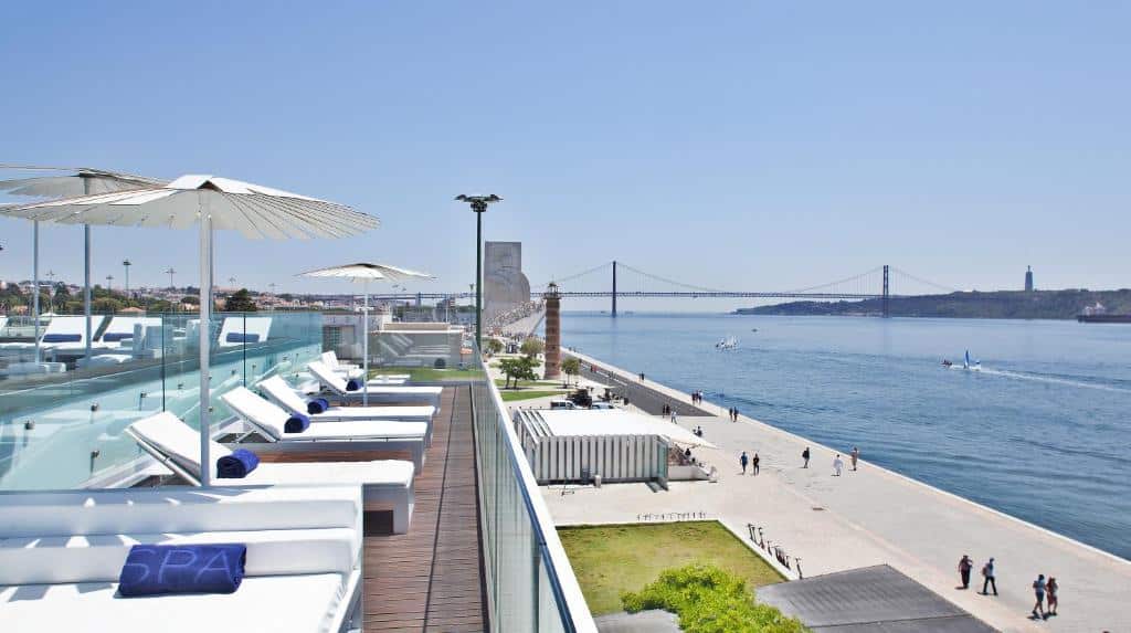 Terraço do Altis Belem Hotel & Spa – Design Hotels com vista para o Rio Tejo, trata-se de um deck onde há uma piscina, no local você vê espreguiçadeiras e guarda-sóis abertos