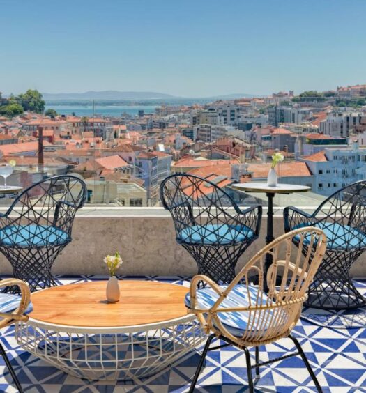 Terraço na cobertura do H10 Duque de Loule com vista para a cidade e, mais ao fundo, dá para ver o rio Tejo, e no terraço há algumas cadeiras e pequenas mesinhas, para representar hotéis bem localizados em Lisboa