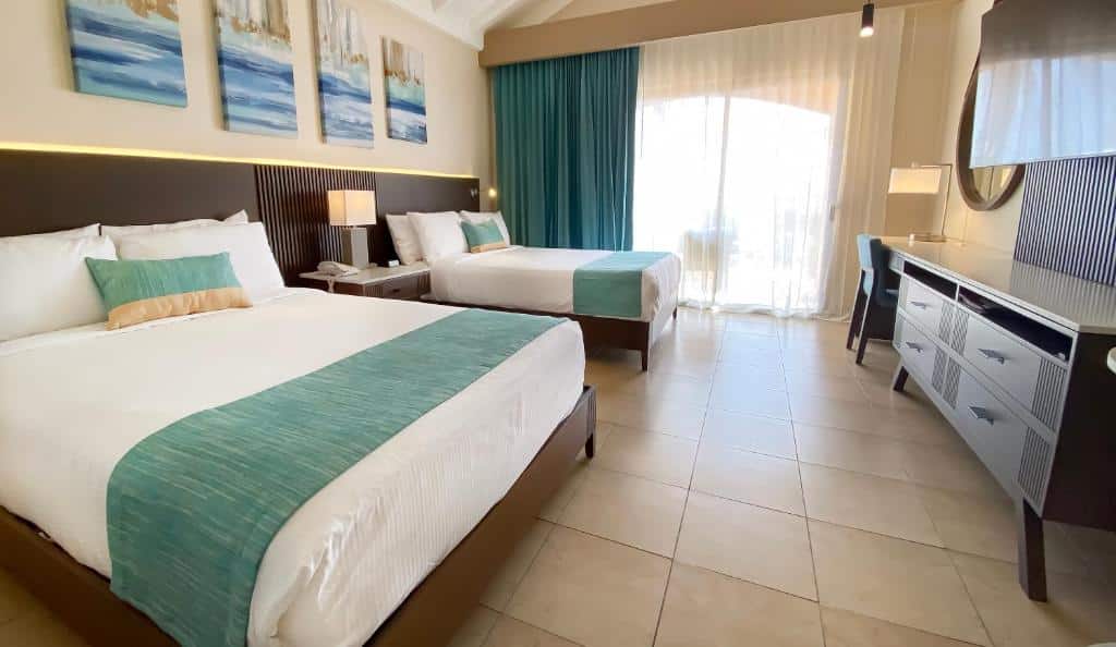 Quarto do The Royal Sea Aquarium Resort. Do lado direito uma mesa de trabalho com abajur, espelho e TV. Do lado esquerdo duas camas de casal, entre elas uma prateleira com abajur. No fundo a cortina fechando a varanda.