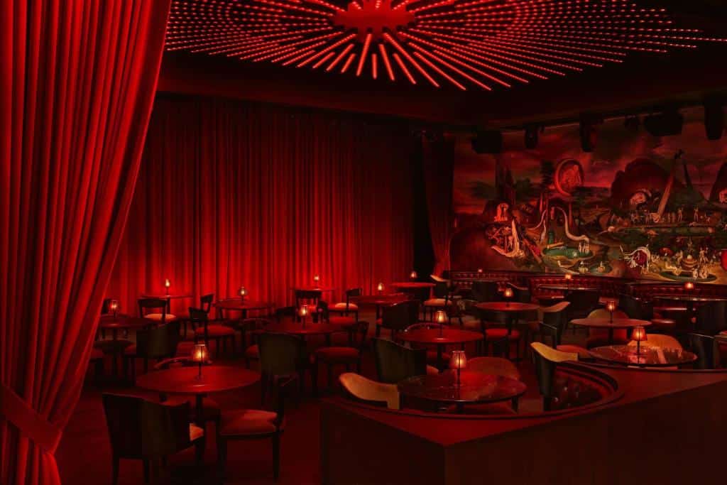 Salão de festas do The Times Square EDITION New York, o local é escuro com luzes indiretas na cor vermelha, há um palco, com mesas e cadeiras de madeira, além de alguns sofás e uma das paredes é inteira desenhada