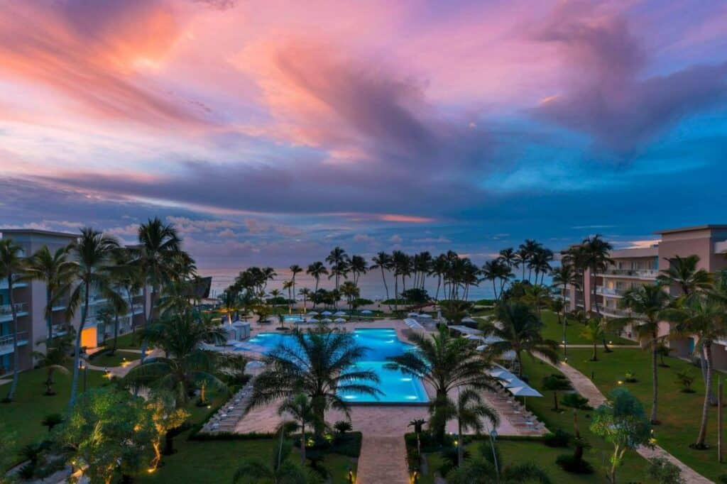 Vista aérea do The Westin Puntacana Resort ao pôr do sol. Uma grande piscina fica no centro e é rodeada por palmeiras. Os prédios do resort ficam nas laterais da imagem, e a praia está ao fundo.