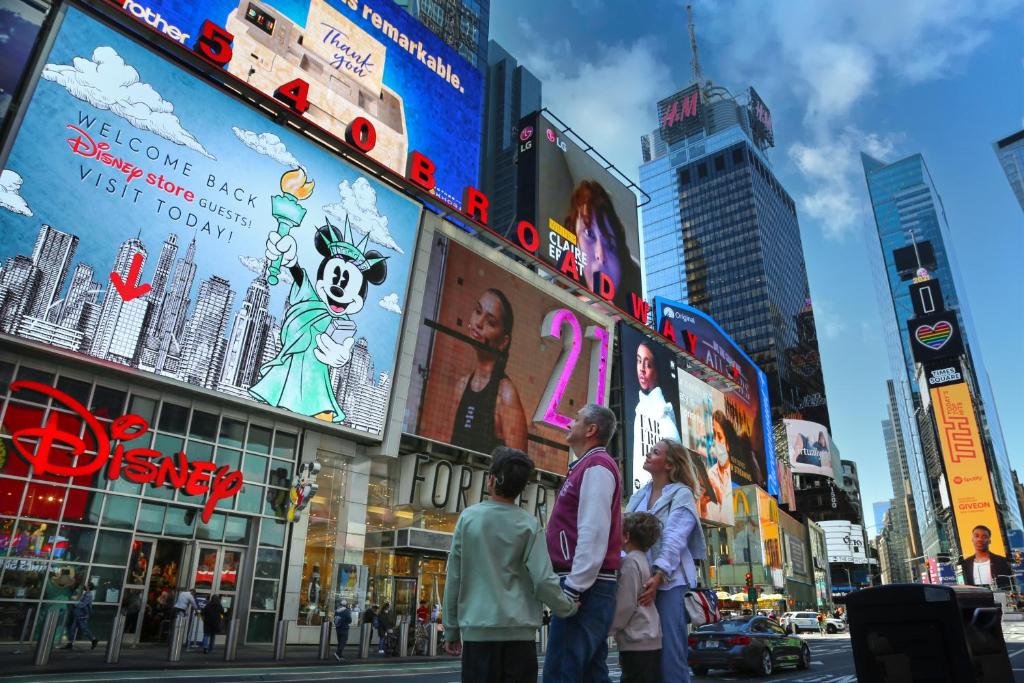 uma família com dois adultos, um homem branco de uns 40 ou 50 anos, dando a mão para um menino pequeno com problema de audição, e uma mulher branca de uns 40 anos, abraçada a um menino pequeno, enquanto todos olham para os outdoors da Times Square em Manhattan.
