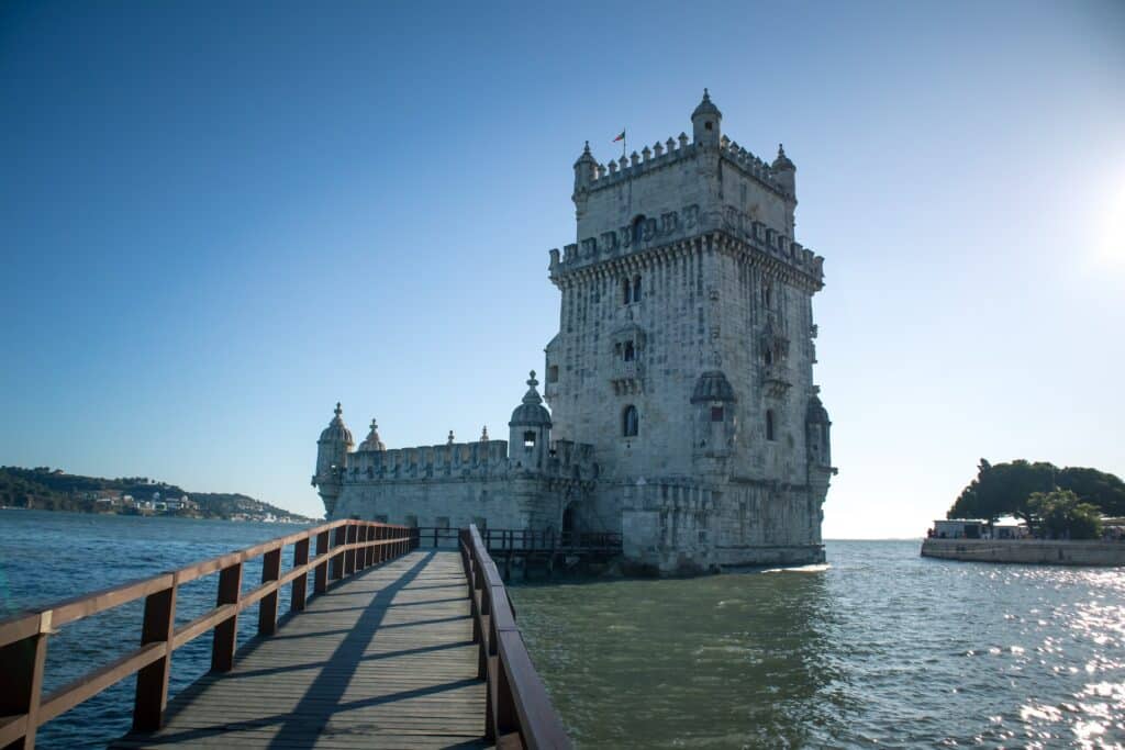 Uma torre de época alta com algumas pequenas janelas, há pequenas torres no topo junto com uma bandeira de Portugal, e há uma ponte de madeira por cima do rio para chegar até a torre