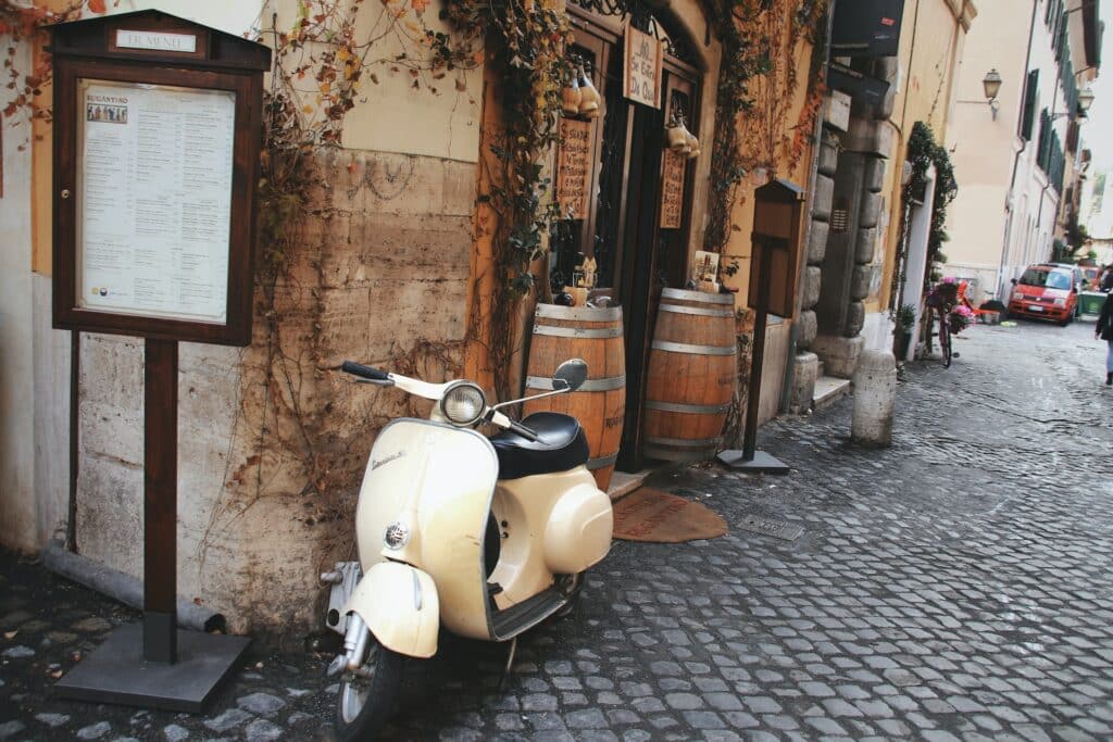 vista de um rua no Trastevere, uma das opções de o que fazer em Roma, com uma vespa branca, barris decorativos e ruas de pedra charmosas