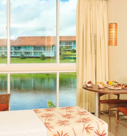 Quarto da Villa Hípica Resort. A cama está no lado esquerdo do quarto, enquanto há uma mesa com café da manhã no lado direito e uma janela com vista para um lago ao fundo. Imagem para ilustrar o post hotéis e pousadas em Gravatá.