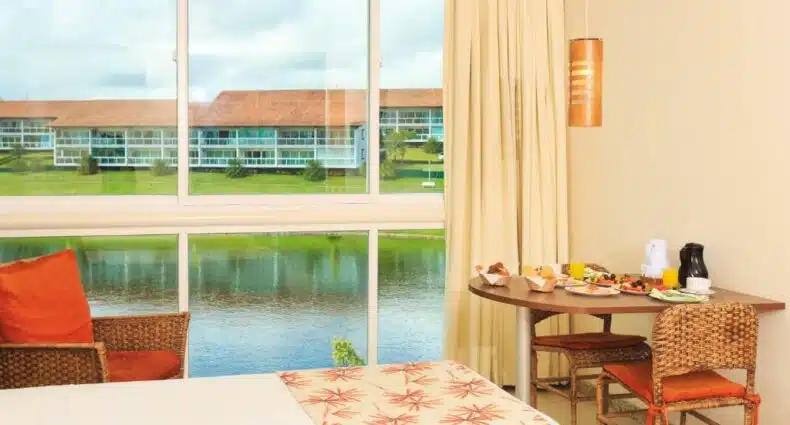 Quarto da Villa Hípica Resort. A cama está no lado esquerdo do quarto, enquanto há uma mesa com café da manhã no lado direito e uma janela com vista para um lago ao fundo. Imagem para ilustrar o post hotéis e pousadas em Gravatá.