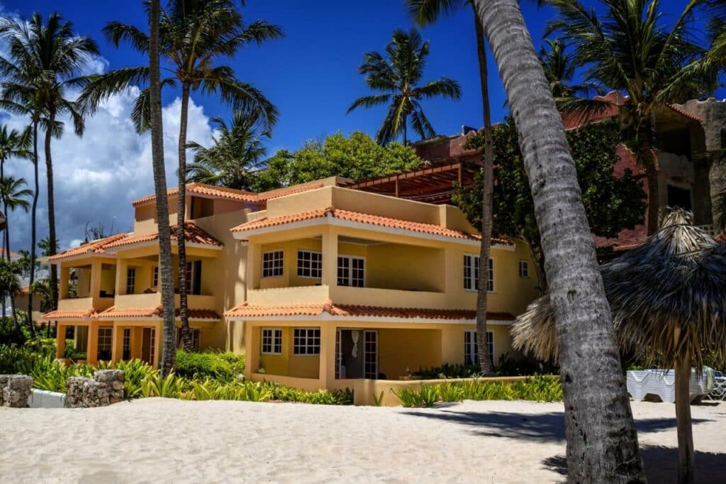Foto externa das Villas Chiara Punta Cana. Uma construção de dois andares amarela na beira da praia tem várias janelas brancas de vidro. Alguns coqueiros e outras plantas estão espalhadas ao redor do local.