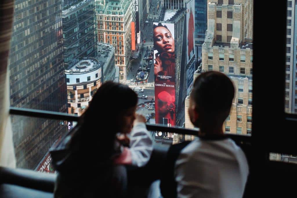 ampla janela com vista para as ruas e os prédios da Times Square. Em frente as janeas há duas pessoas olhando para fora, uma mulher e um rapaz de cabelo curto.
