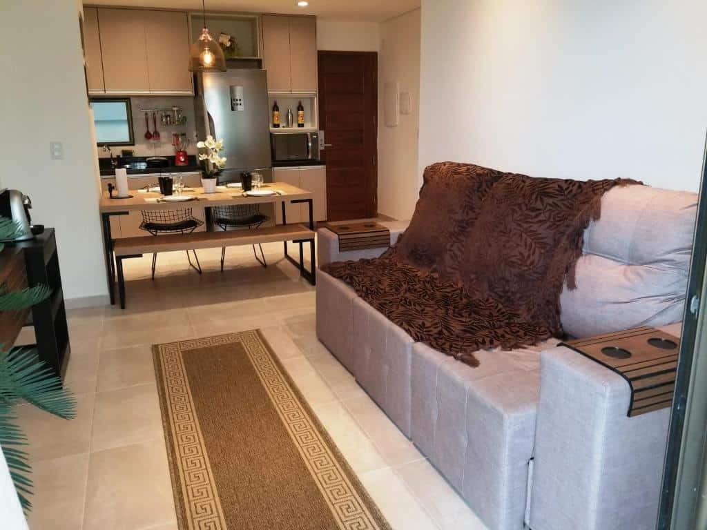 Parte do Wai Wai Cumbuco Eco Residence - Bahamas 101 que mostra um pedaço da sala de estar com um sofá retrátil, um tapete no chão e ao fundo uma mesa de madeira com dua cadeiras e uma cozinha completa com geladeira, microondas e uma pia.