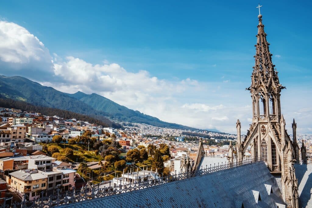 vista de Quito a partir da Basílica do Voto Nacional, com uma torre com muitos detalhes, várias casinhas e uma montanha atrás com céu azul e nuvens