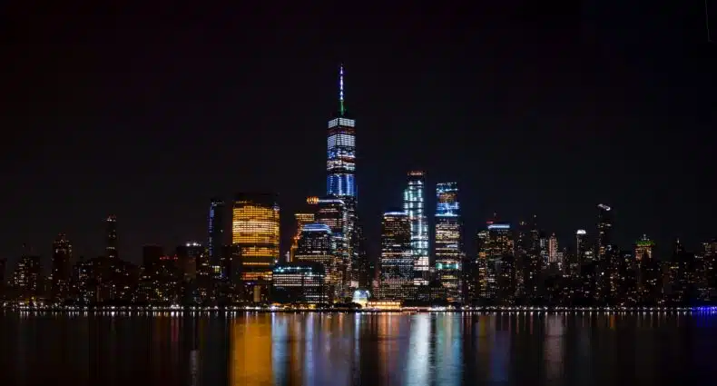 vista à noite do Wall St., Financial District, em Nova York, com prédios luminosos com seus reflexos na água