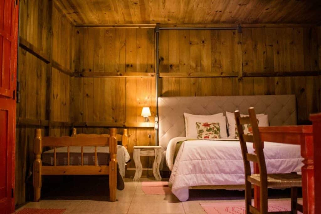 Quarto da Afeto Pousada, em Bento Gonçalves, com uma cama de casal, com lençóis brancos e cabeceira estofada, uma mesinha de cabeceira com luz de leitura sobre ela, e uma cama de solteiro ao lado