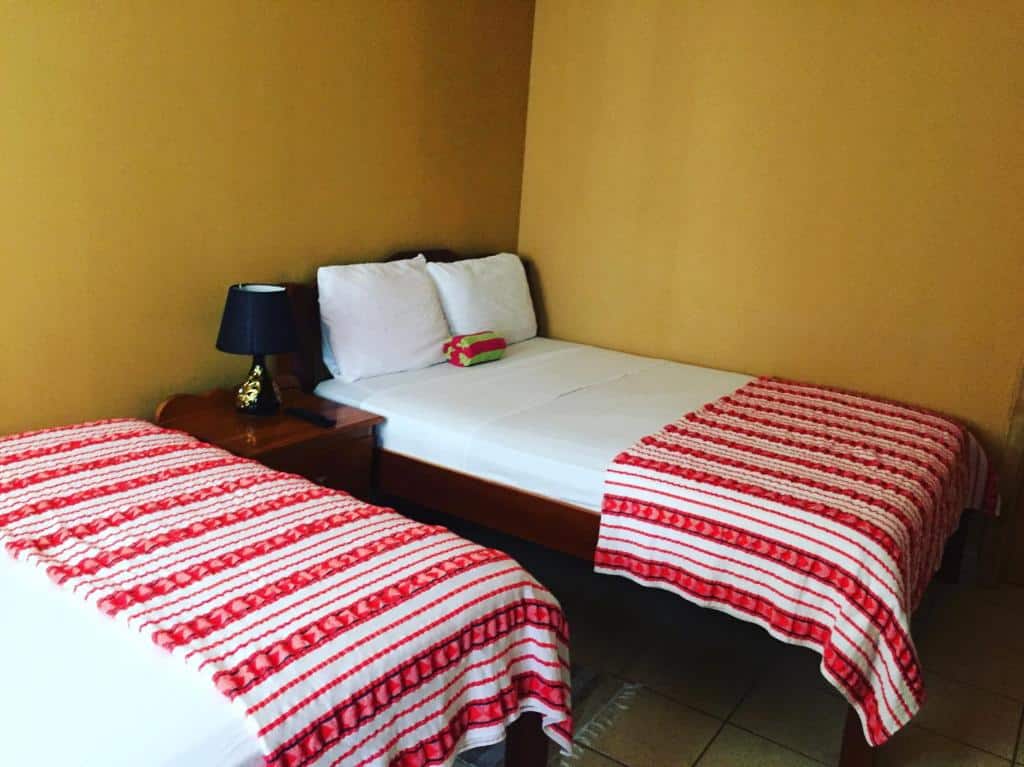 Um quarto no Ambergris Sunset Hotel, para representar o post sobre hotéis em Belize. Há duas camas de casal. Uma delas pode ser vista parcialmente no canto inferior esquerdo da foto. A outra está de frente para a primeira, encostada na parede do fundo. Há uma mesa de cabeceira com abajur em cima no seu lado esquerdo.
