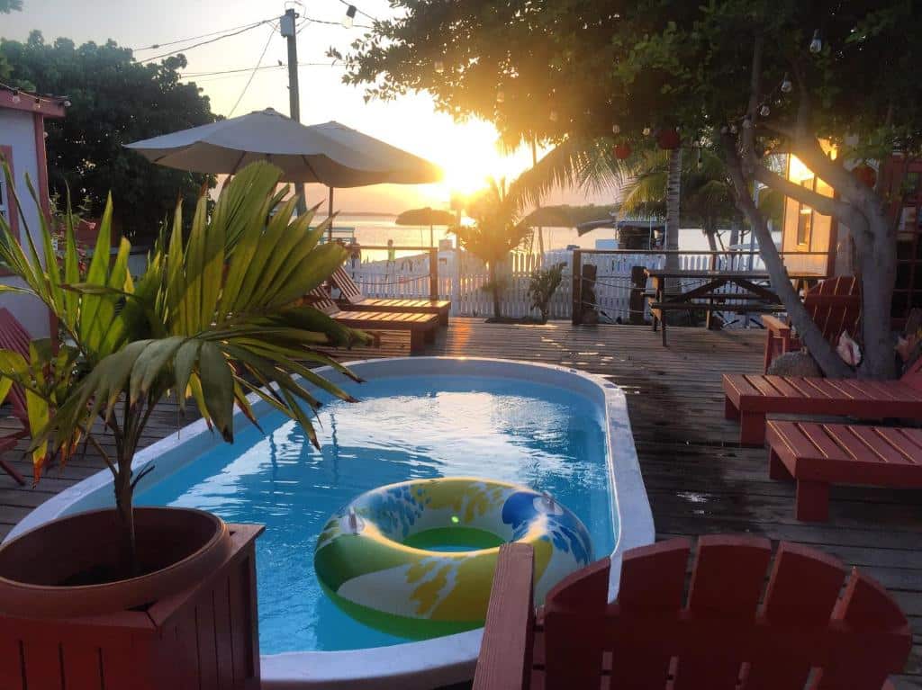 Área externa do Ambergris Sunset Hotel. Vemos um deck com piscina, essa que possui uma boia dentro, espreguiçadeiras, vasos com plantas e um guarda-sol. No fundo vemos o sol se pôr no horizonte da praia.