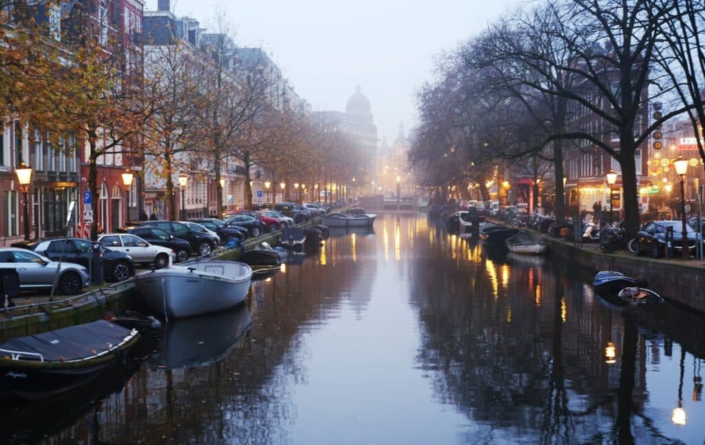 Canal de Amsterdam, um dos destinos mais visitados da Europa, com barquinhos parados nas margens, e ruas em ambos os lados, onde há carros estacionados, árvores secas e prédios coloridos