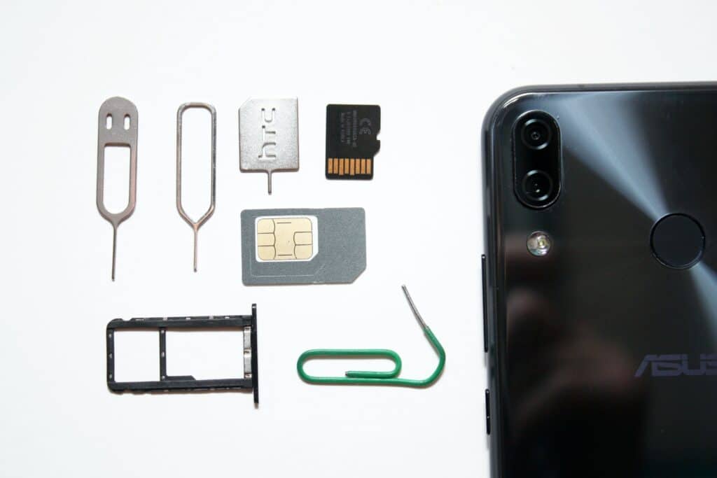 celular Asus deitado sobre uma mesa branca, com a tela para baixo. Ao lado esquerdo há vários itens de chip, como um cartão SIM, a gaveta de chip do celular, a chave de abertura e até o cartão de memória.
