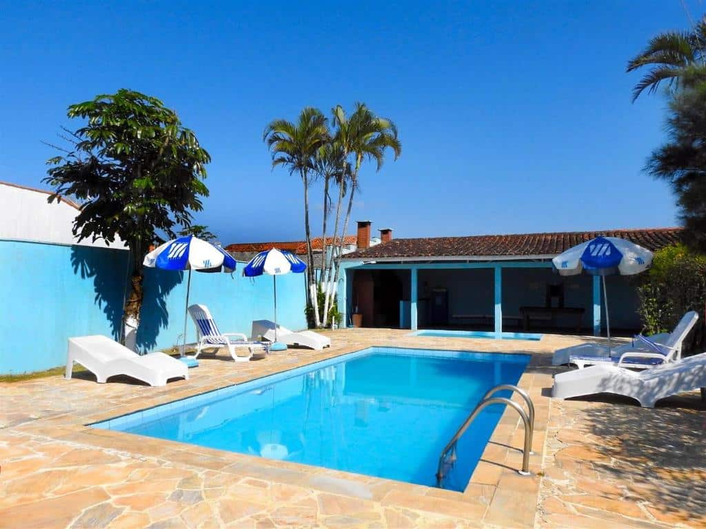 Piscina ao ar livre da Associação Sabesp Ilha Comprida. No lado direito uma escada para entrar na piscina, ao redor cadeiras de tomar sol e guarda-sóis. No fundo, outra piscina pequena e um espaço com churrasqueiras.