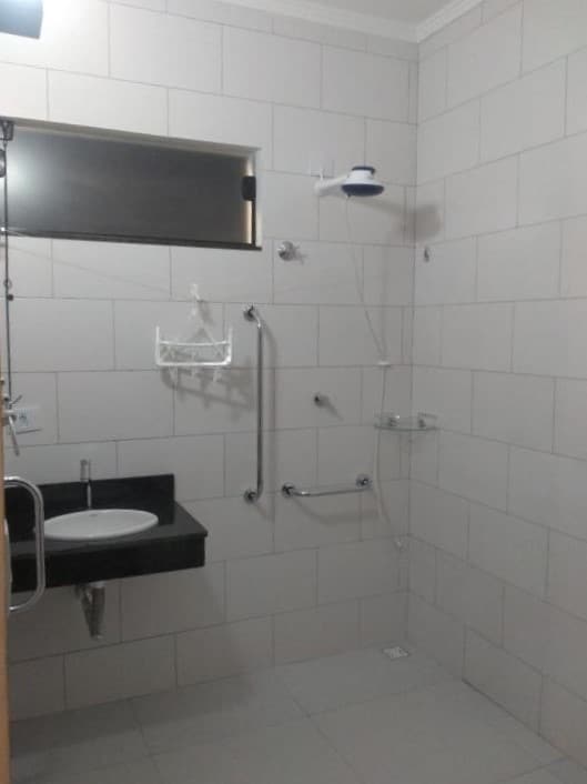 Banheiro comm acessibilidade do Balli Suítes. Uma pia no lado esquerdo, no lado direito o chuveiro com barras de apoio.