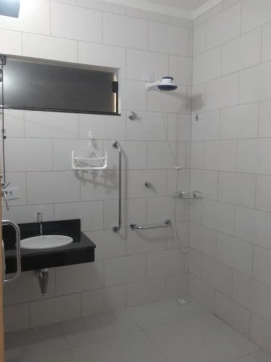 Banheiro comm acessibilidade do Balli Suítes. Uma pia no lado esquerdo, no lado direito o chuveiro com barras de apoio.