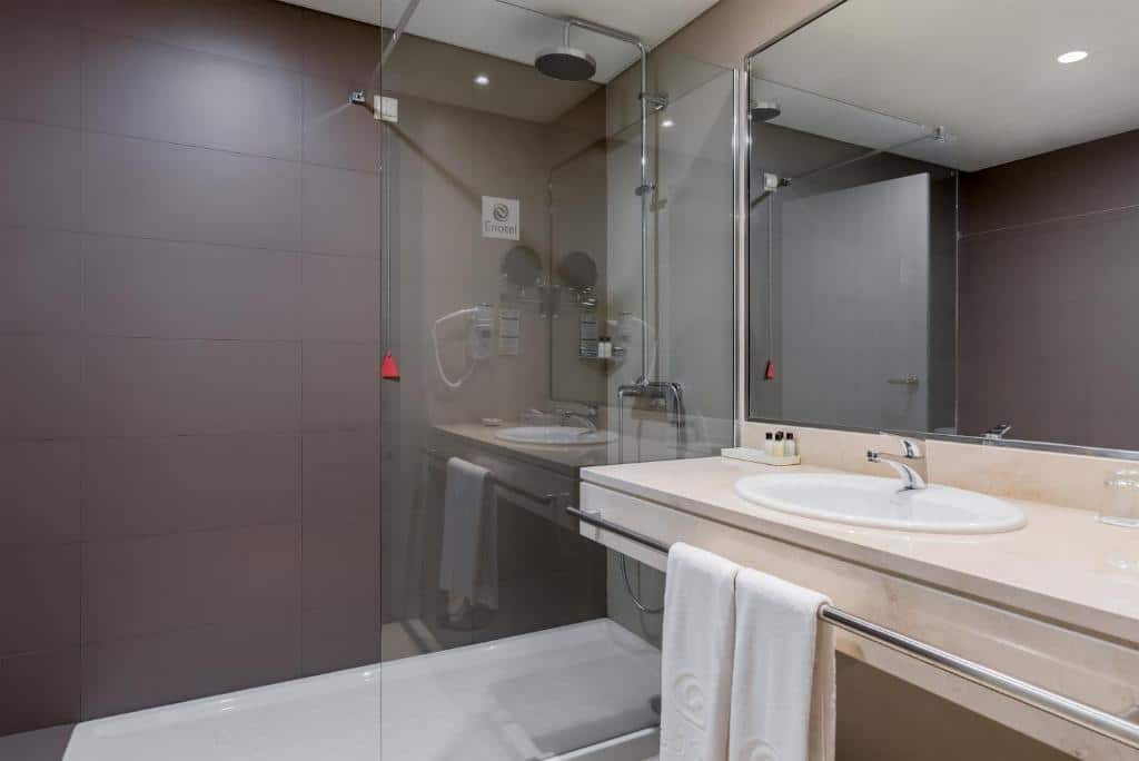 Banheiro com acessibilidade do Enotel Lido – All Inclusive com pia baixa com barra de apoio do lado direito da imagem ao fundo box de vidro com banheira. Representa hotéis all inclusive em Portugal.