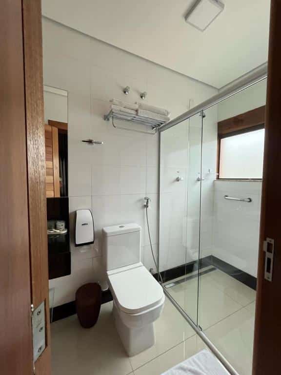 Banheiro na Pousada mar Mineiro Macacos. No centro, há uma privada branca com ducha, e do lado, vemos parcialmente uma bancada preta de mármore que tem uma pia. Do outro lado da privada, está o box, que é espaçoso e possui ducha, uma barra de apoio e janela.