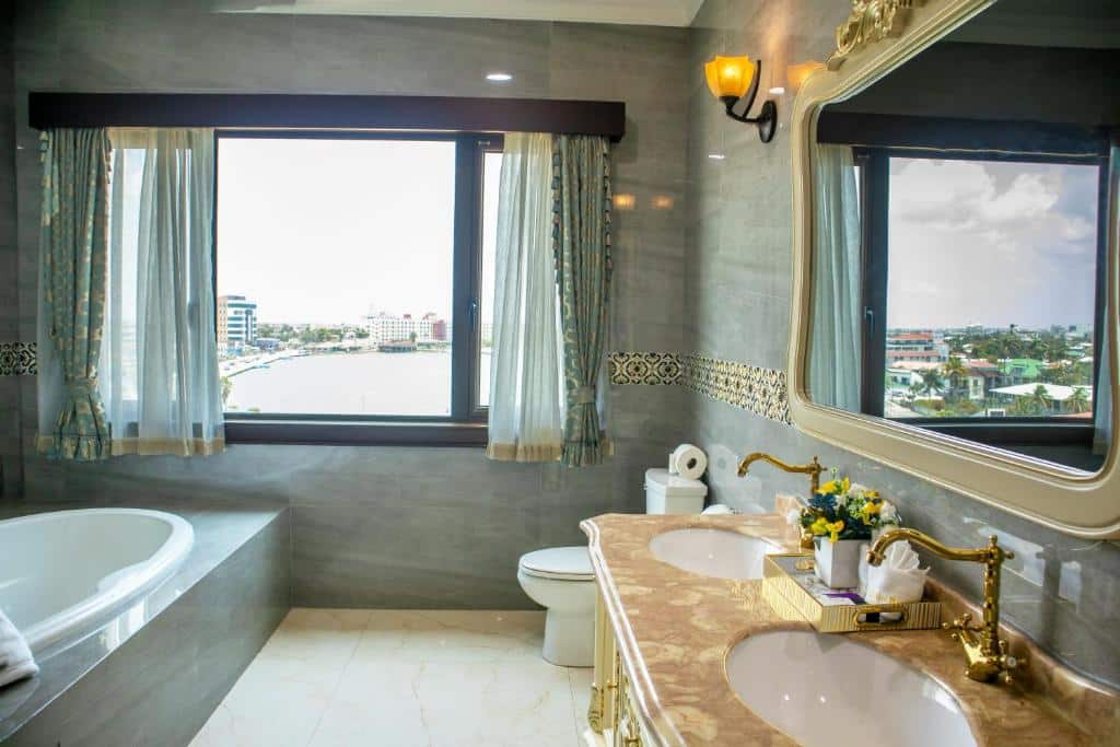 Banheiro do Golden Bay Belize Hotel. No canto inferior esquerdo podemos ver de relance uma banheira, e na sua frente há uma janela. Já no canto inferior direito está a cabine da pia, que tem detalhes clássicos. Ela possui duas pias, uma bandeja com amenidades em cima e um espelho à sua frente. Do lado esquerdo da pia, no fundo do ambiente, há um vaso sanitário.