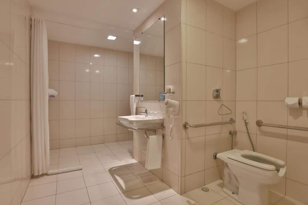 Banheiro do Quality Hotel & Suítes São Salvador. Ele é acessível, possui um vaso sanitário rebaixado no canto direito da imagem com grades de apoio. Uma pia rebaixada está mais ao centro do ambiente, que também tem barras de apoio e um espelho largo. No fundo, está a ducha com uma área bastante larga para banho, com cortinas para privacidade. O banheiro em geral é bastante espaçoso.