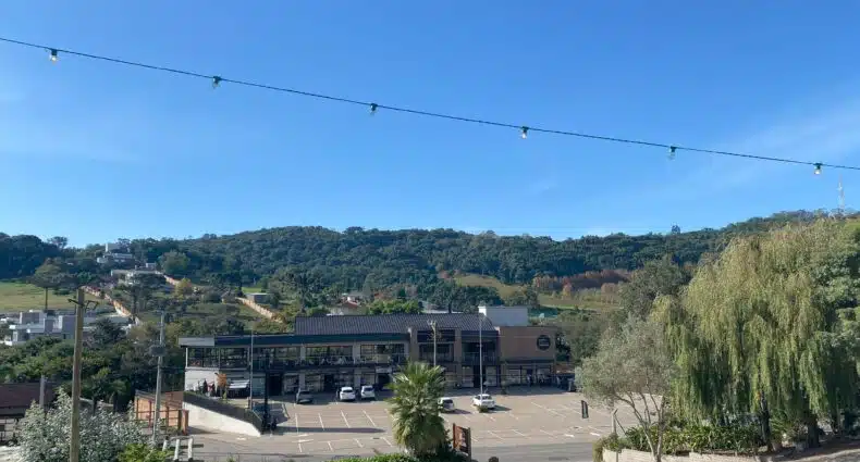 Vista de Bento Gonaçalves, no RS, com shopping a céu aberto cheio de lojinhas, cafés e restaurantes, em dia de céu azul com nuvens esparsas e escassas, montanhas cheias de árvores ao fundo, e algumas árvores nos cantos, mais próximos da tela