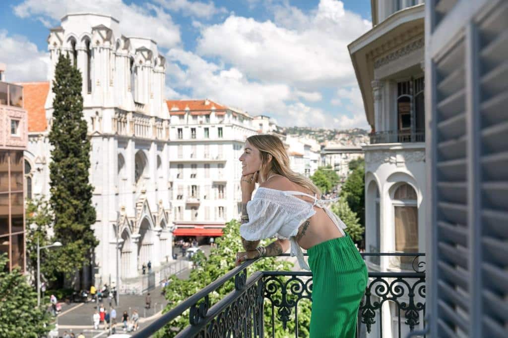 Varanda do Best Western Hotel Lakmi Nice. Na imagem há uma mulher apoiada no parapeito da varanda sorrindo e admirando a vista das ruas de Nice, onde há diversas construções elegantes.