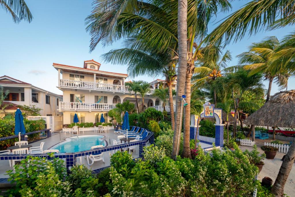 Foto externa do Blue Tang Inn. Há várias árvores e arbustos. No canto inferior direito é possível ver um quiosque. Do lado dele, há uma entrada para a área da piscina do hotel. Essa área possui muitos azulejos brancos e azuis, e espreguiçadeiras e guarda-sóis na mesma cor. A piscina pode ser vista no lado esquerdo da imagem. No fundo está o hotel, e podemos ver suas varandas e janelas.