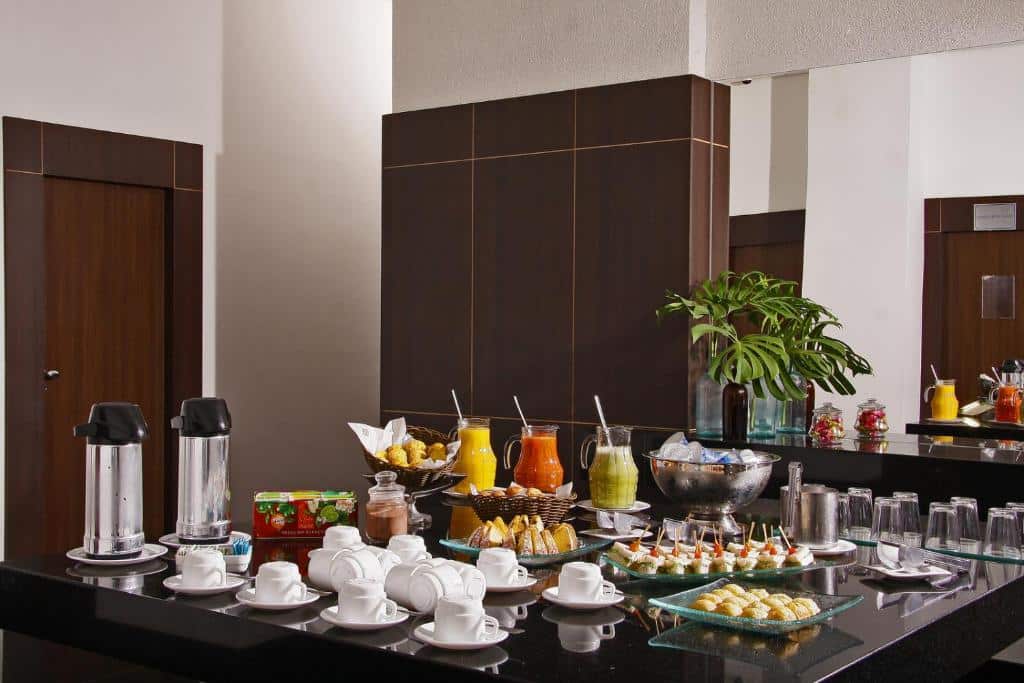 Um buffet de café da manhã no restaurante do Piemonte Hotel. Vemos uma grande mesa de mármore preta dispondo várias xícaras brancas, jarras de suco e de café, copos de vidro, doces, pães, bolos, etc.