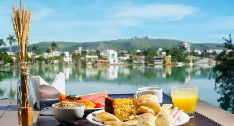 Foto de um café da manhã ao ar livre no Hotel Atlas. Em cima de uma mesa, vemos um prato com uma fatia de bolo, pão francês, salgados, frios, e pães de queijo. Na frente do prato, há um copo com suco e uma xícara de café ao lado. Também há outro prato com frutas, uma tigela com cereais e um suporte com guardanapos. Ao fundo da imagem, vemos um grande lago, prédios e montanhas.