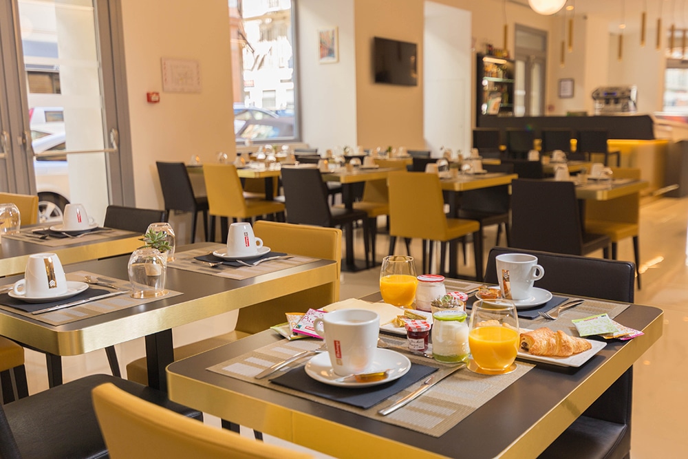 Área de refeições do Hôtel Nice Azur Riviera. Na mesa há xícaras de café, copos de suco, iogurte e croissant. No fundo há diversas outras mesas.