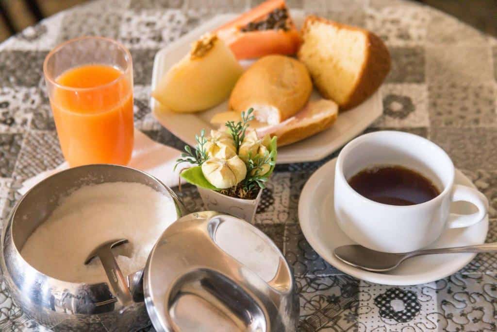 Uma foto do café da manhã na Pousada Rio Branco Nova Lima. Podemos ver uma xícara branca com café na direita, um açucareiro ao seu lado, mais para a esquerda da foto, um copo com suco de laranja atrás e um prato branco com pães, frutas e uma fatia de bolo.