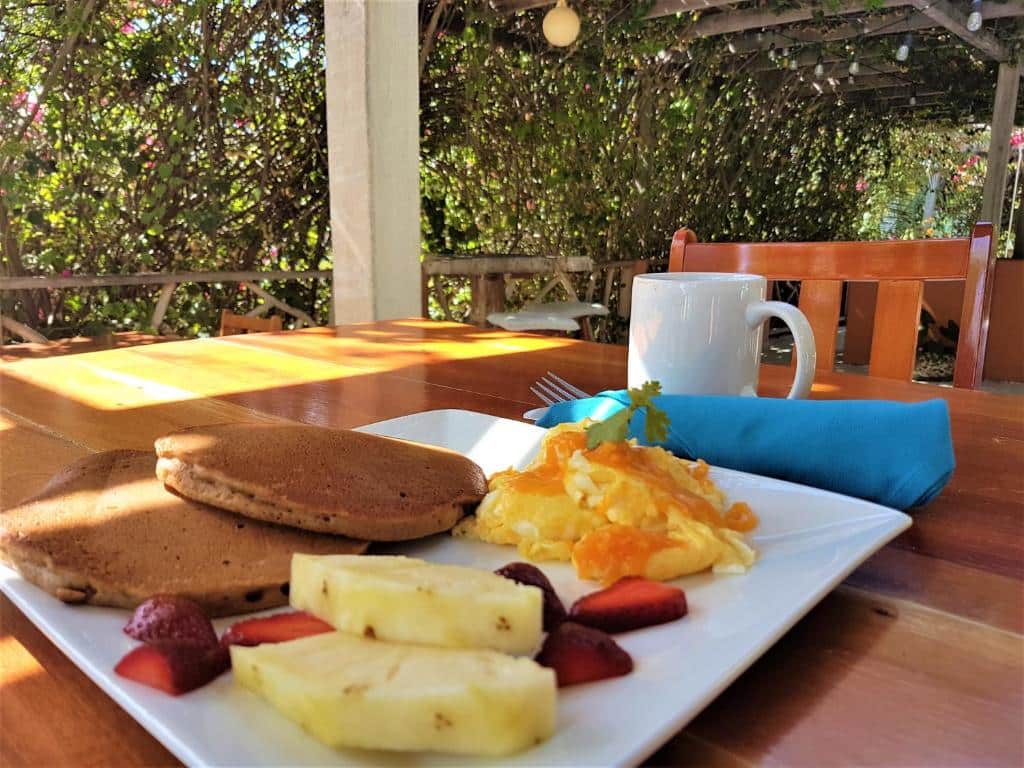Um café da manhã ao ar livre no Sea Dreams Hotel. Vemos um prato com frutas e panquecas em cima de uma mesa de madeira. Também há um guardanapo azul e uma xícara branca. Há uma cadeira do outro lado. No fundo, vemos muitas árvores.