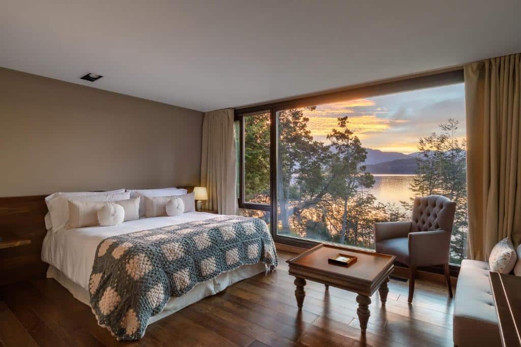 Quarto do Calfuco Wine Hotel & Spa. Uma cama de casal no lado esquerdo, de frente uma mesinha, um sofá e uma poltrona. Uma parede atrás com vista para o lago.