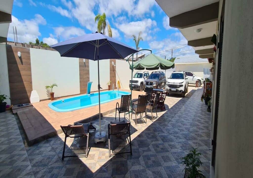 Área da piscina da Cananeia Suítes. A piscina está no canto direito, embaixo do sol. Ao seu lado esquerdo estão espreguiçadeiras e dois guarda-sóis. Ao fundo estão quatro carros estacionados, para representar pousadas em Cananéia.