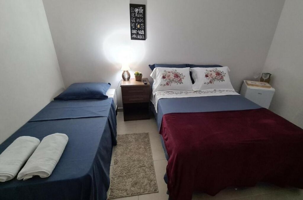 Quarto da Cananeia Suítes. Existem duas camas lado a lado, uma de casal e outra de solteiro. No meio delas há uma pequena mesinha com abajur.