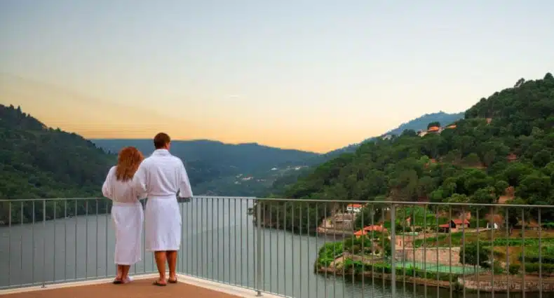 Casal parado na varanda no Douro Royal Valley Hotel & Spa, durante o dia olhando o rio Douro. Representa hotéis para lua de mel em Portugal.