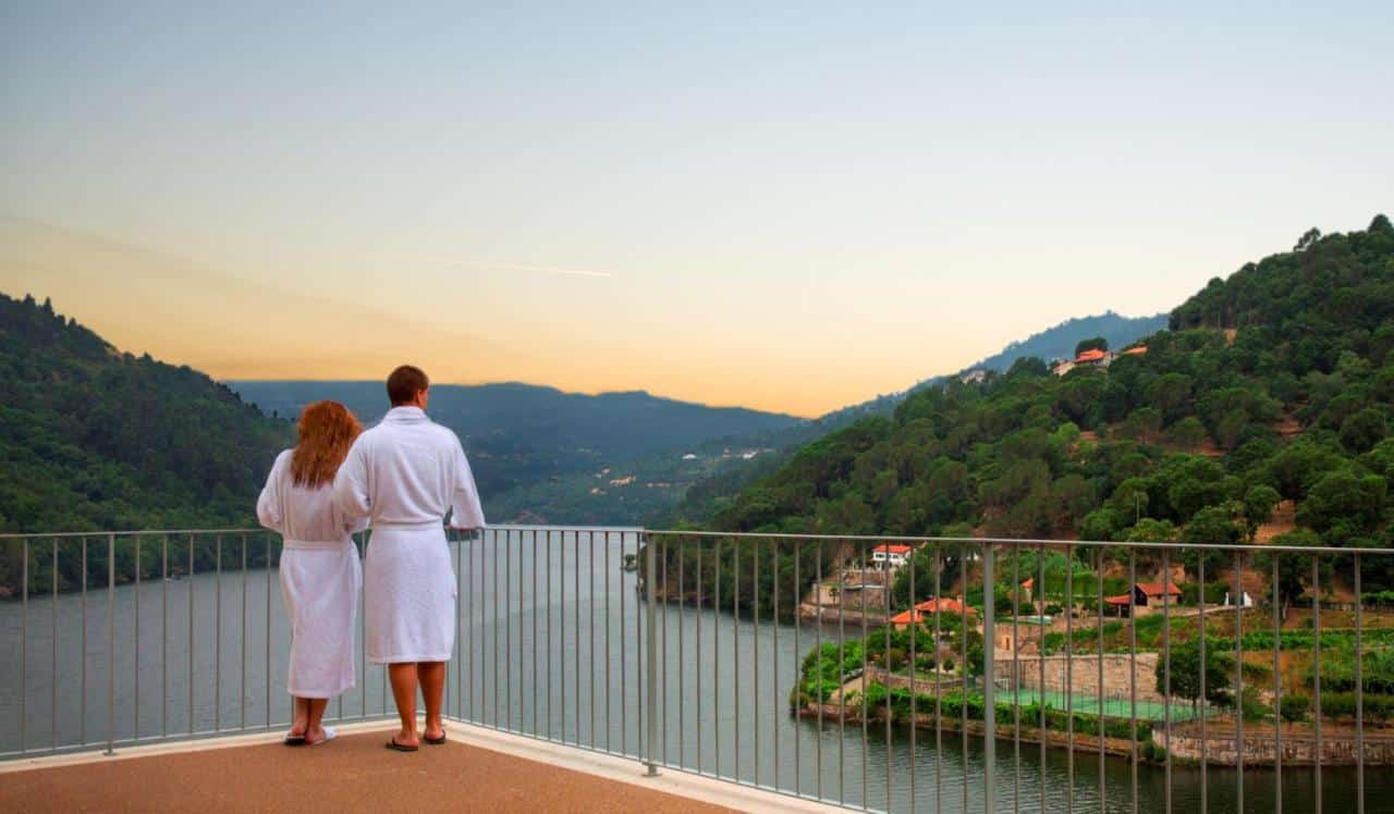 Casal parado na varanda no Douro Royal Valley Hotel & Spa, durante o dia olhando o rio Douro. Representa hotéis para lua de mel em Portugal.
