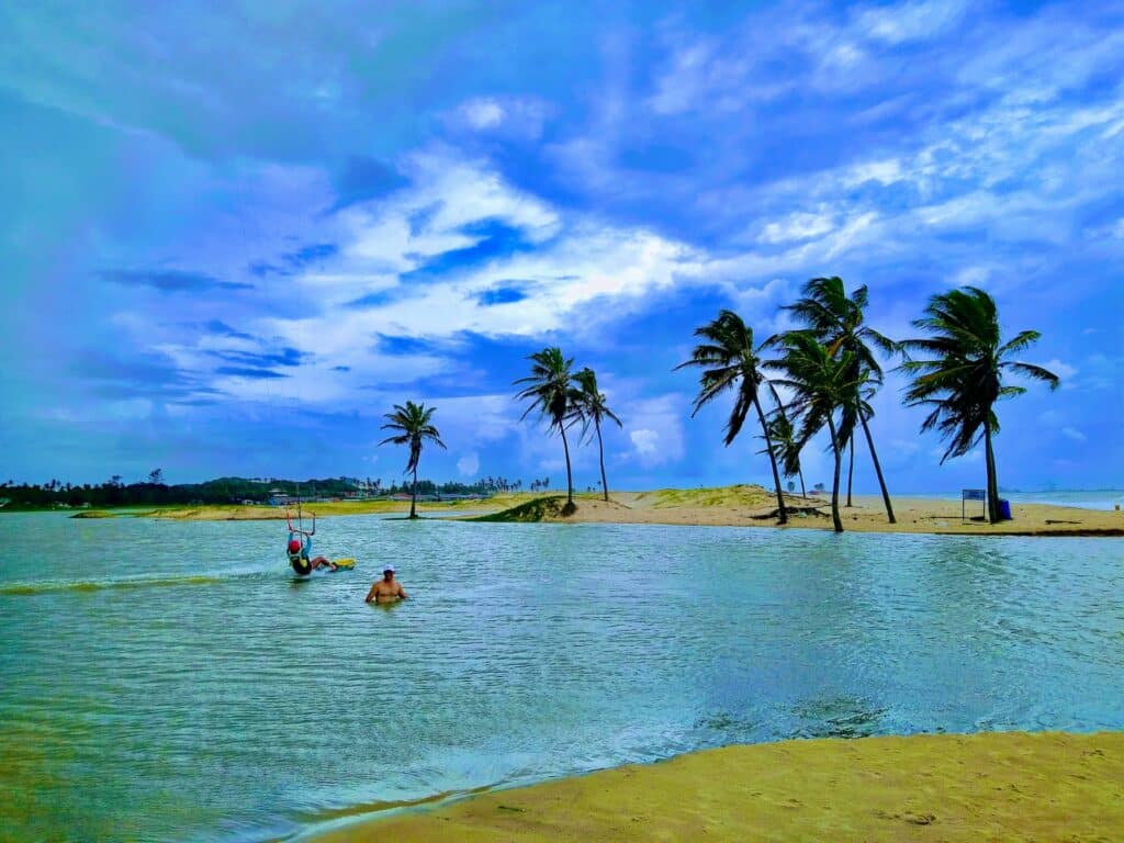 Lagoa do Cauipe Cumbuco com uma pessoa na água e a outra praticando kitesurf logo atrás. Ao fundo tem algumas palmeiras e dunas de areia durante o dia, ilustrando post Hotéis em Cumbuco.