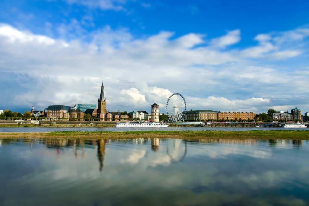 Vista da cidade de Düsseldorf, Alemanha durante o dia com rio a frente e um barco navegando no rio, ao fundo a cidade com construções germânicas e uma roda gigante.