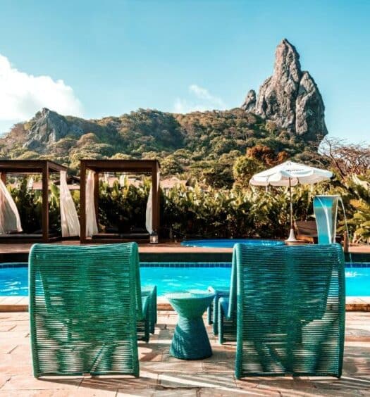 Duas cadeiras de tomar sol na frente, com uma mesinha de apoio entre elas. Atrás a piscina, e depois um pergolado com cortina e almofadas, do lado uma piscina redonda pequena e uma cascata. No fundo, um jardim. Foto do Dolphin Hotel para ilustrar post sobre hotéis em Fernando de Noronha.