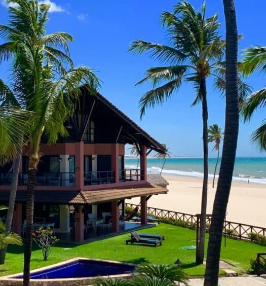 Fachada do Dream Beach Cumbuco Oceanfront Apartments com dois andares de frente para o mar com um gramado em volta, algumas palmeiras e uma pequena piscina azul durante o dia, ilustrando post Hotéis em Cumbuco.