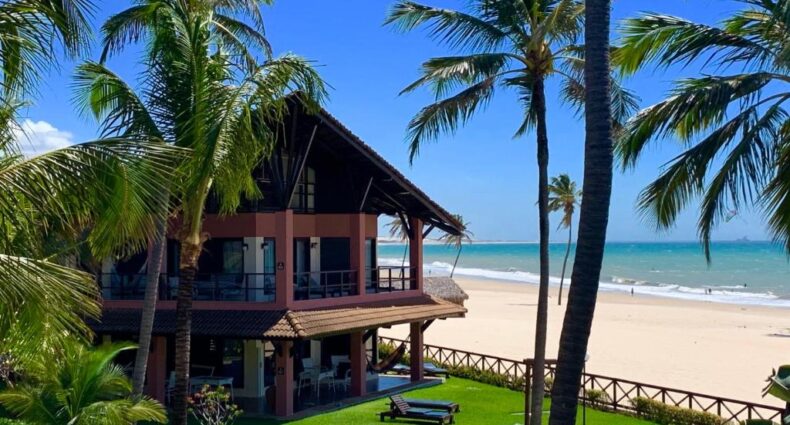 Fachada do Dream Beach Cumbuco Oceanfront Apartments com dois andares de frente para o mar com um gramado em volta, algumas palmeiras e uma pequena piscina azul durante o dia, ilustrando post Hotéis em Cumbuco.