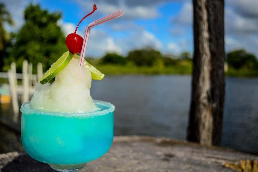 Foto de um drink do Lamanai Hotel & Marina. A taça é de vidro e o conteúdo dentro dela é azul, com bastante gelo, limão, cereja e sal na borda. Possui também um canudo. No fundo, vemos uma paisagem ao ar livre de um rio e árvores.